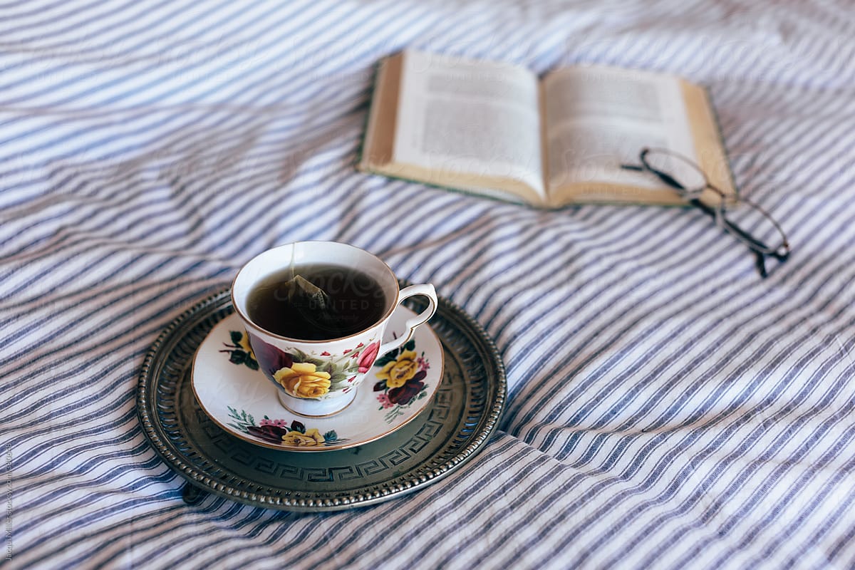 Black tea in china cup on vintage tray on bed, with open book and glasses
