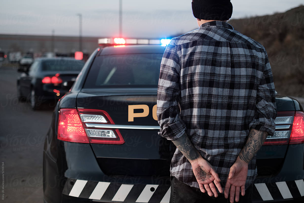 A handcuffed man with tattoos stands at the back of a police cruiser