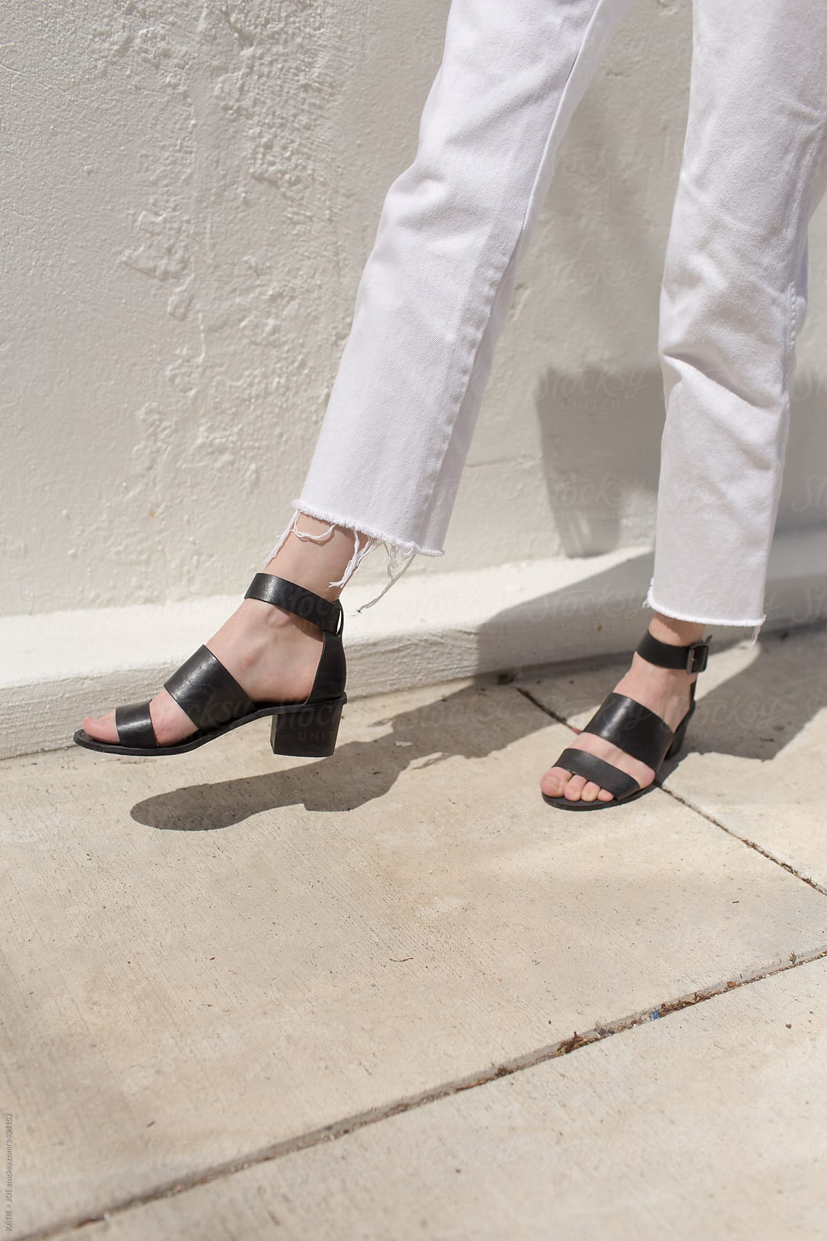 woman\'s legs taking a step on a sidewalk wearing white pants and black sandals