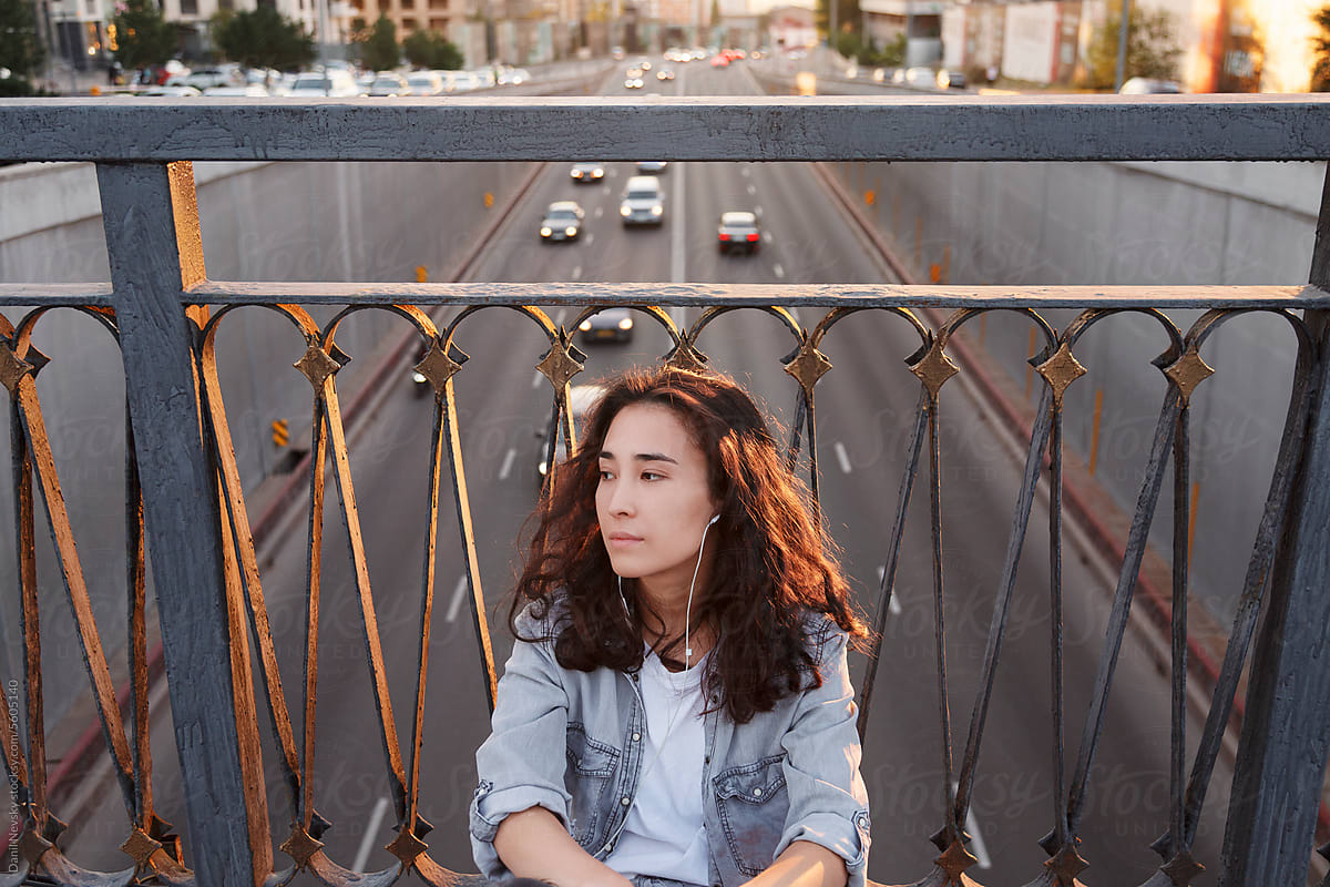 Young woman sitting near metal fence of bridge in day