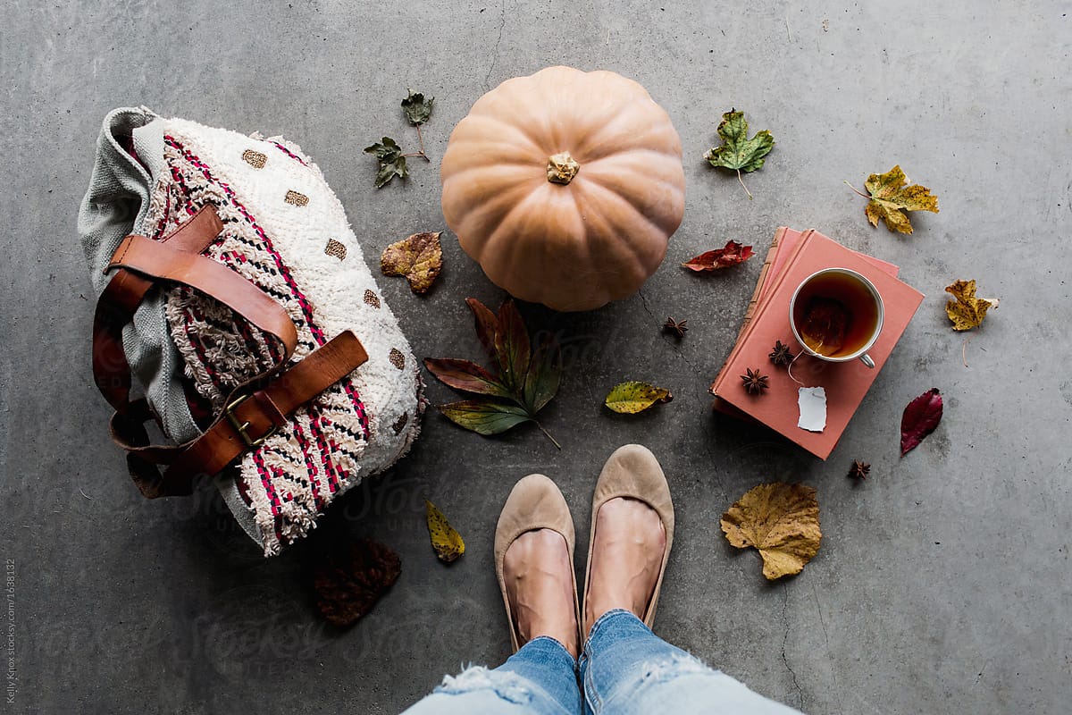 woman in autumn standing next to a pumpkin, books, cup of tea, and a handbag
