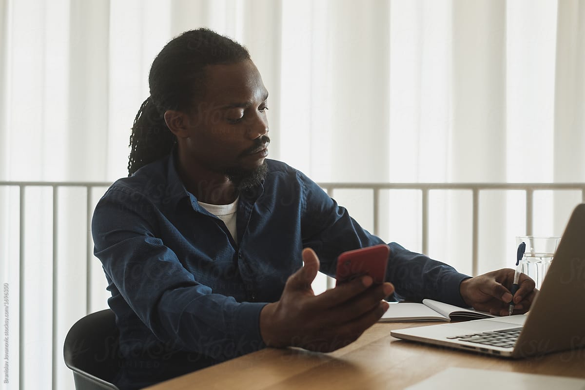 Black Businessman Sitting At The Desk And Using Phone At The Office
