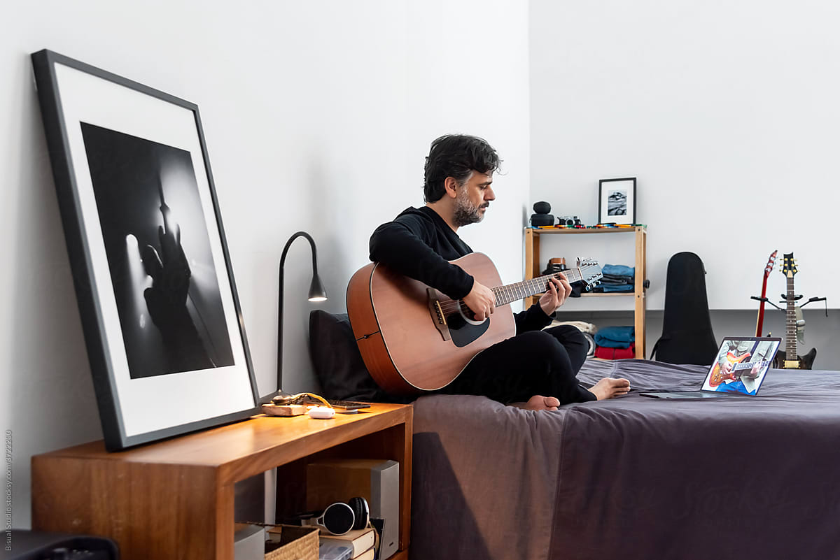Focused man with laptop playing guitar in bedroom