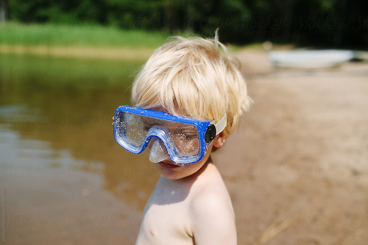 Blonde boy in oversized blue goggles on a beach with a forest in the background.