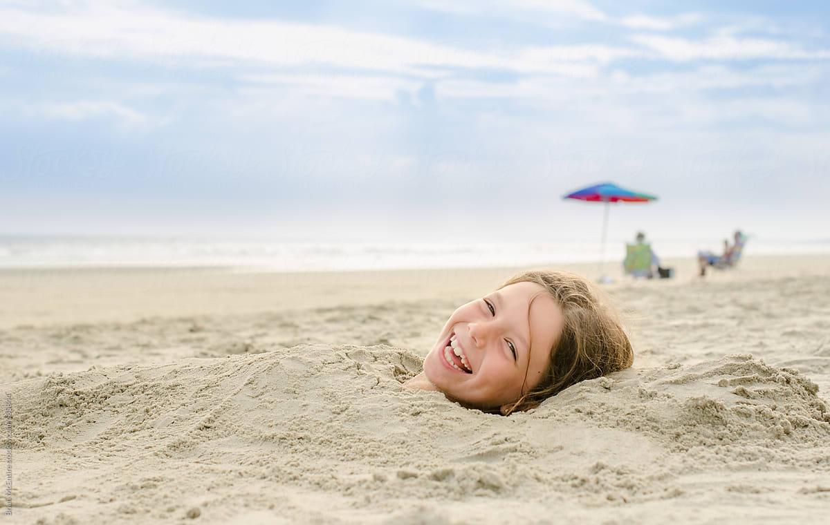 Laughing Girl Buried In Sand At Beach Del Colaborador De Stocksy