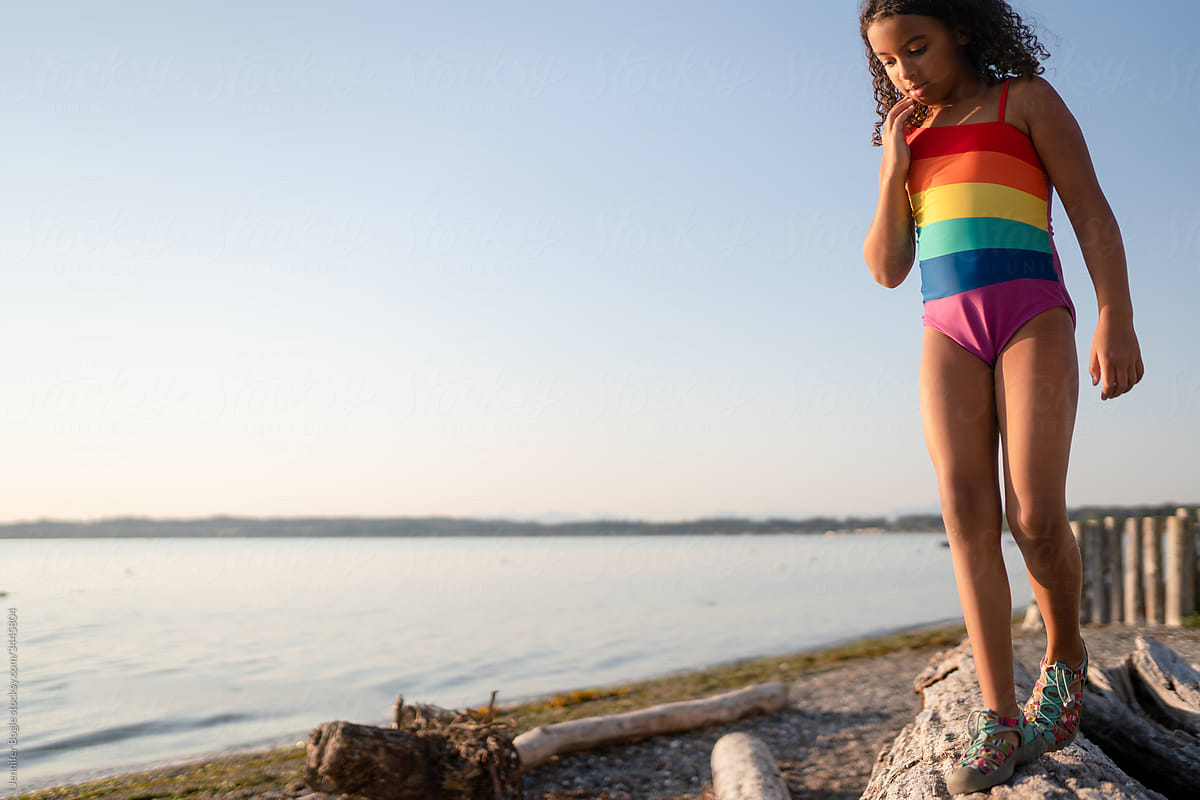 Curly haired girl in rainbow swimsuit walks on log by beach