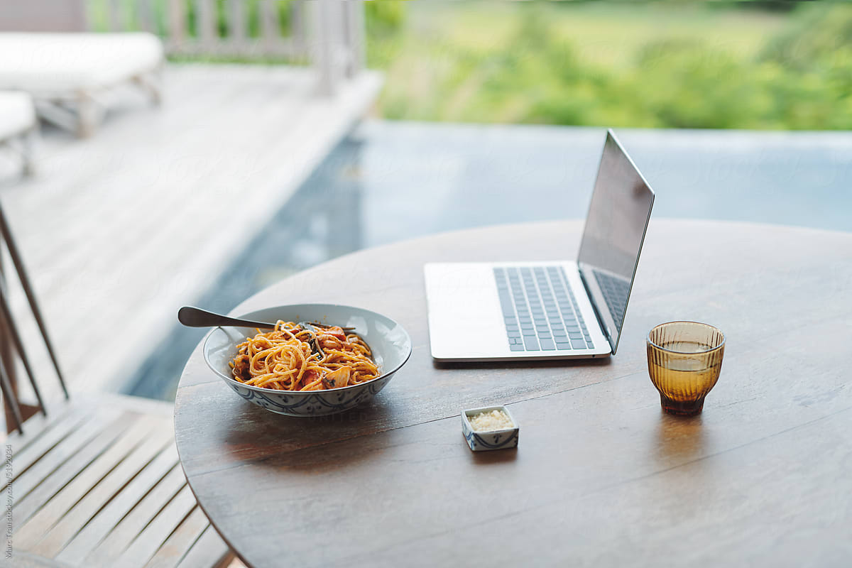 Open laptop and pasta in a plate on wooden table on the patio at home