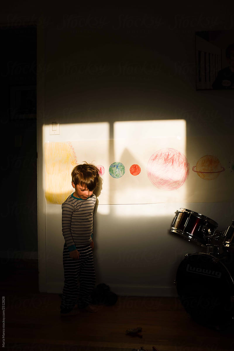 boy stands in sunlight inside with planet drawings behind him and drum set