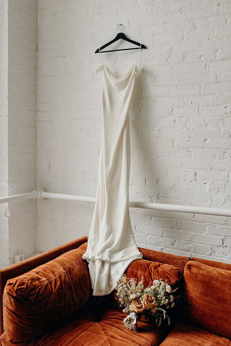 White wedding dress hanging on white brick wall of loft onto burnt orange velvet couch