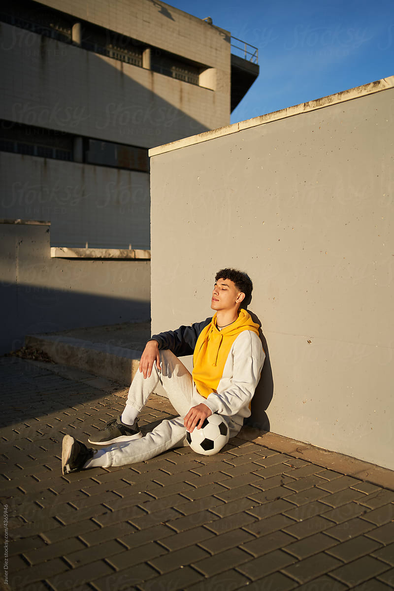Relaxed man with soccer ball sitting near concrete wall