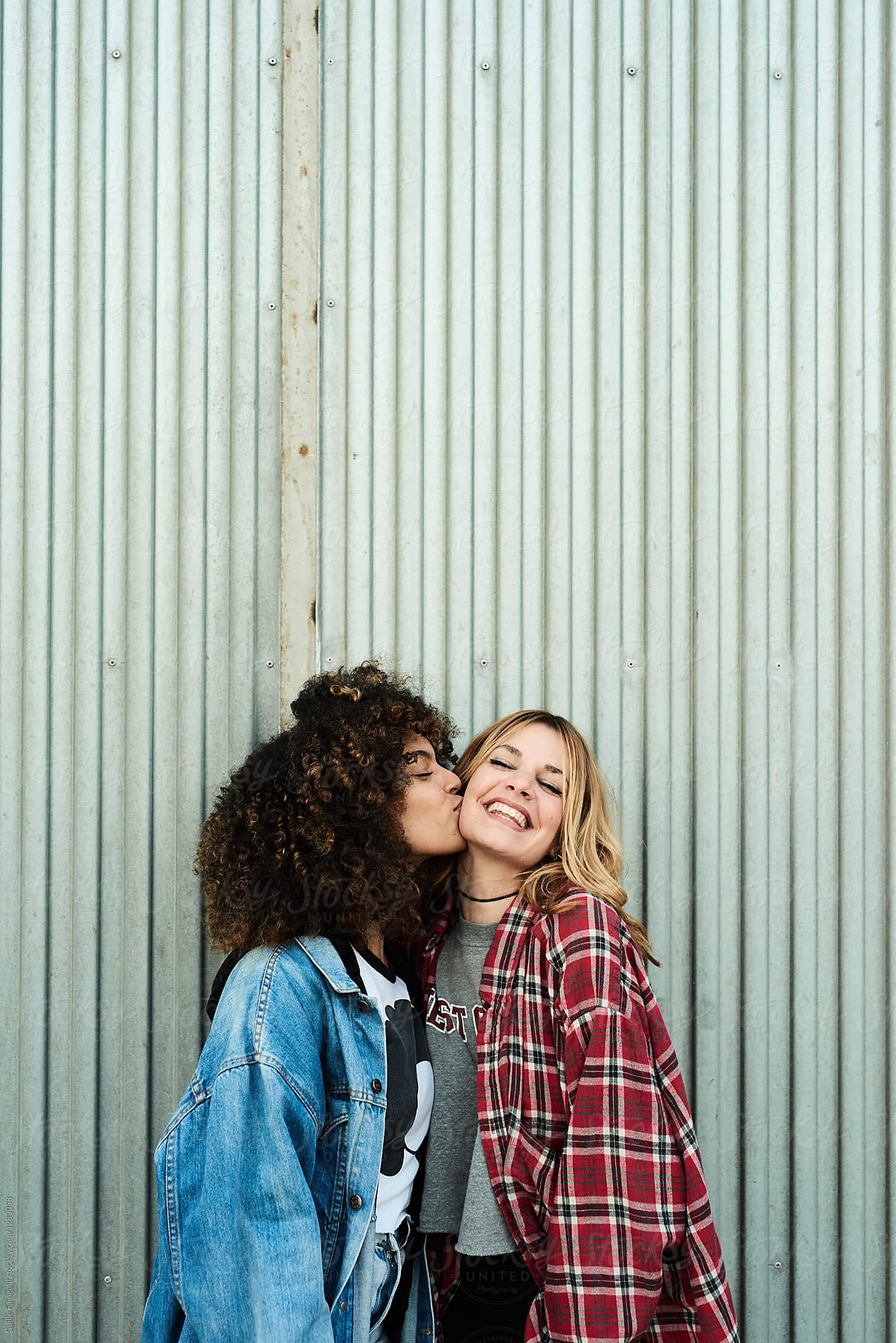 Happy diverse girlfriends kissing against metal wall
