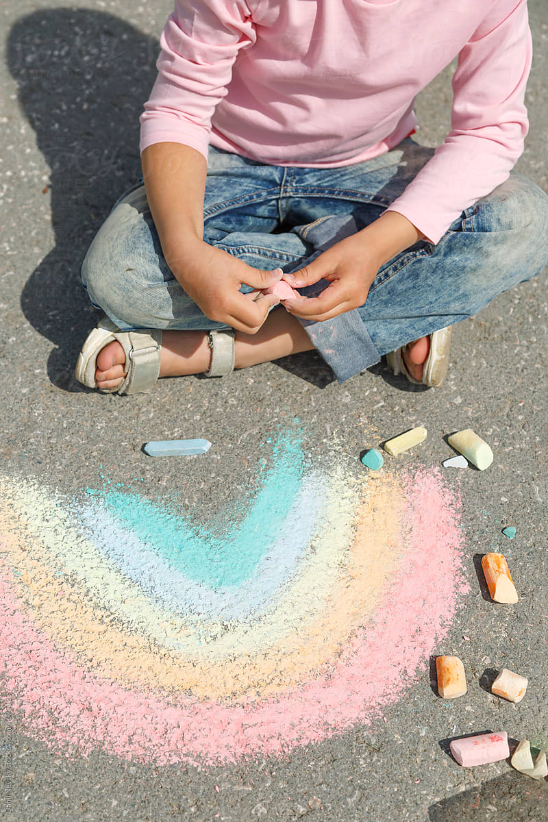 Crop child sitting on asphalt near chalk rainbow