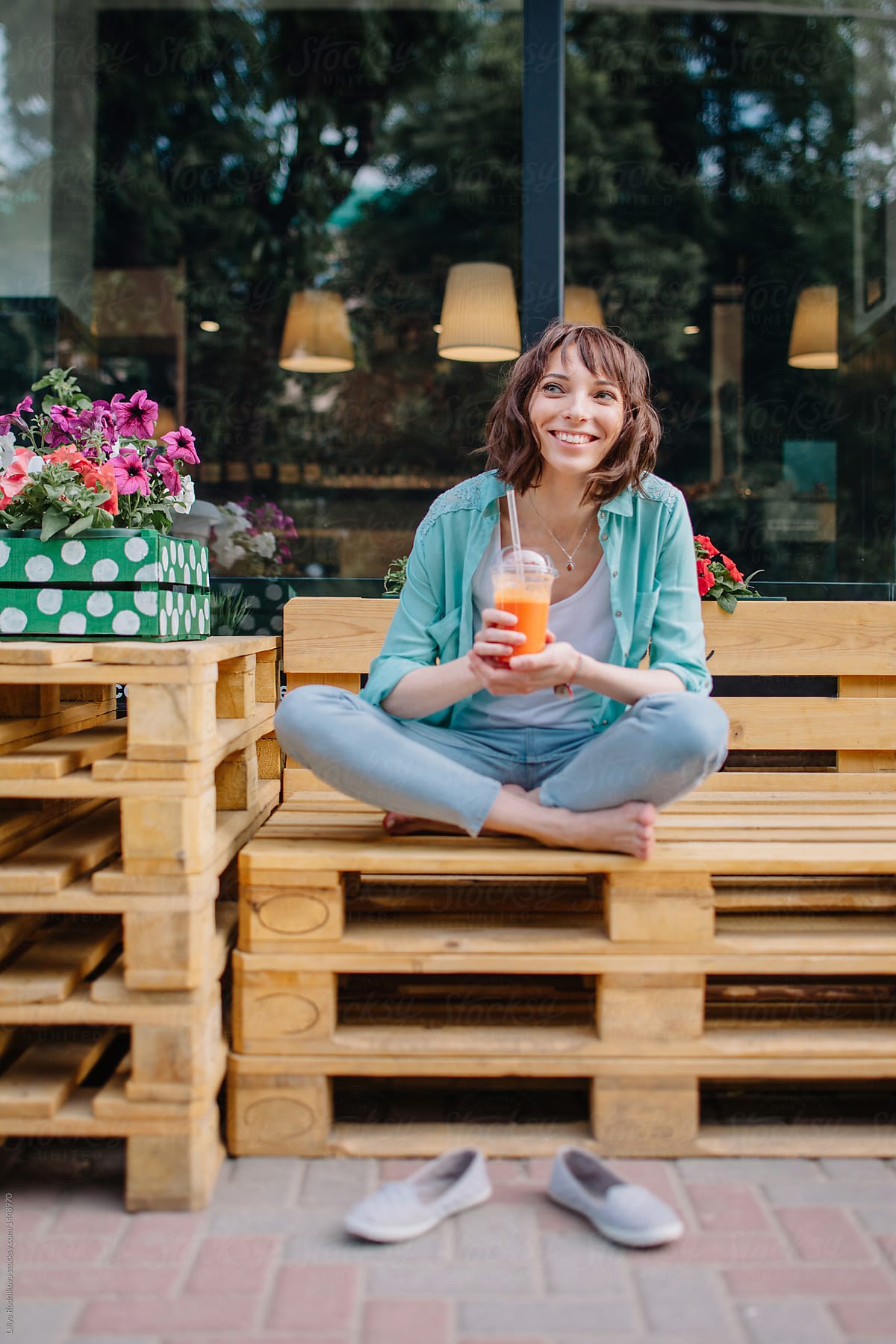 Urban lifestyle shot of smiling woman sitting barefoot on the bench in lotus pose with orange beverage in her hands