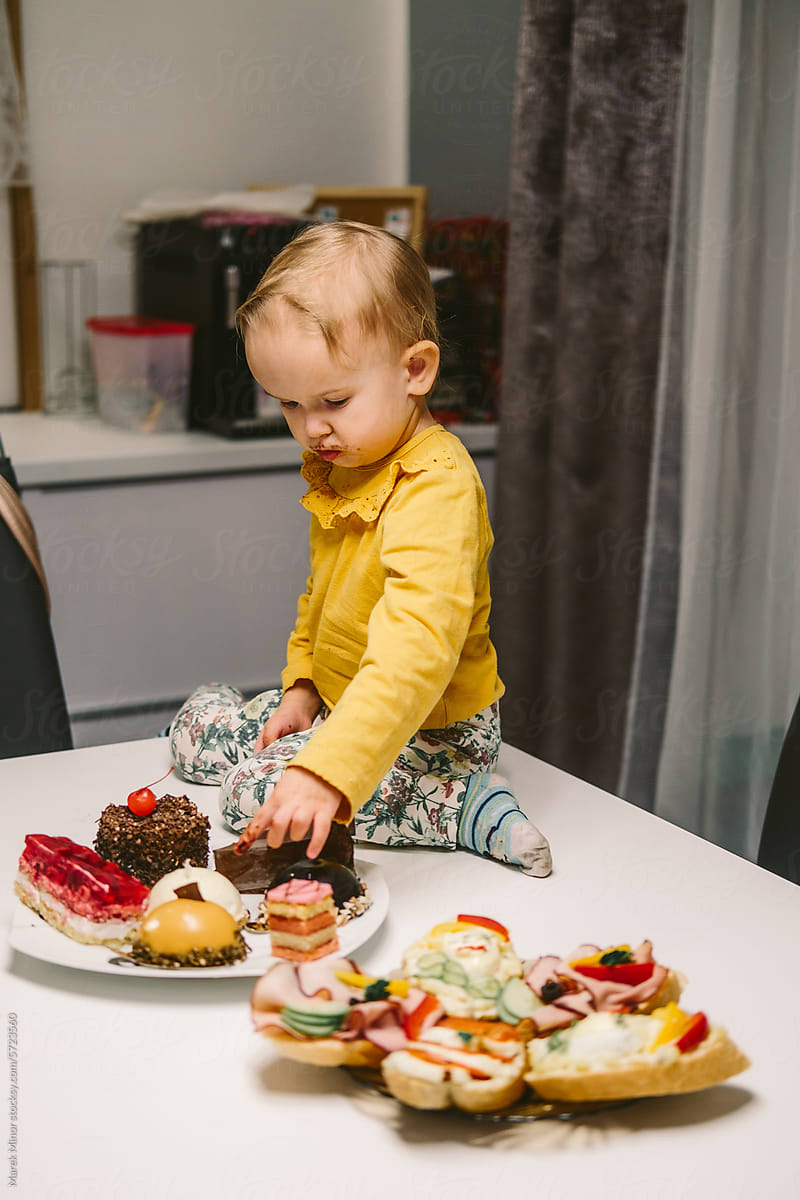 Small baby girl sitting on a table while picking a cake