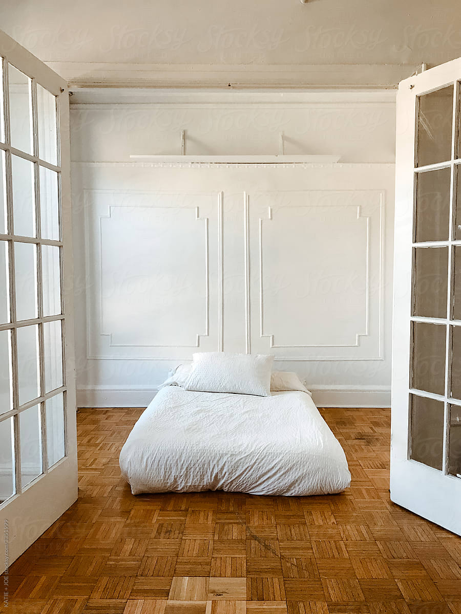 Minimal white bed in an empty apartment with French doors and wooden floors