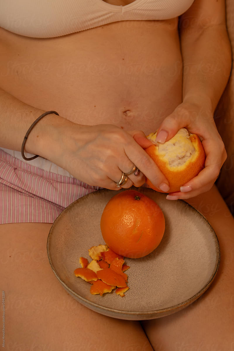 Crop pregnant woman peeling orange