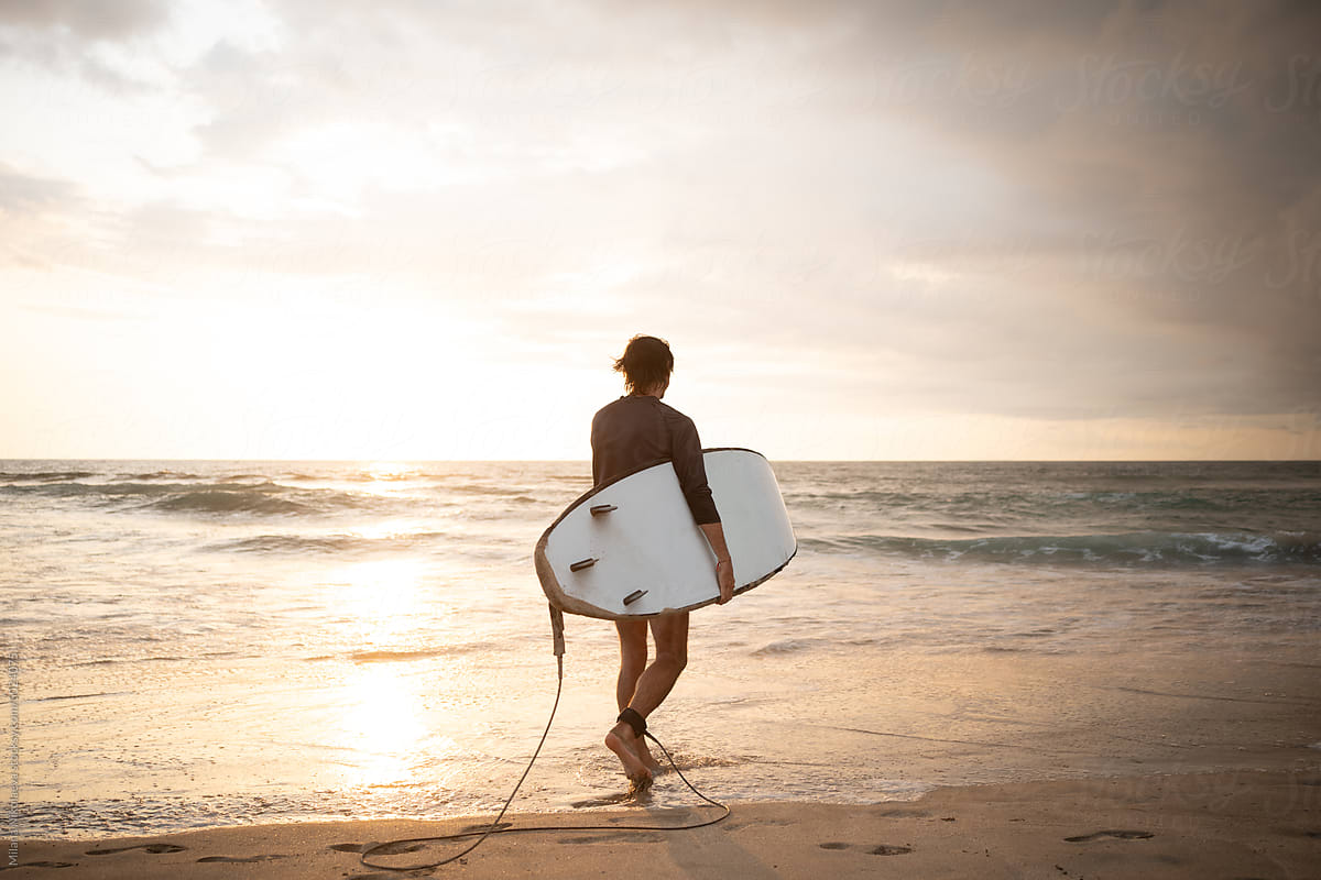 Male Surfer Walking Towards the Waves at a Sandy Beach During Sunset