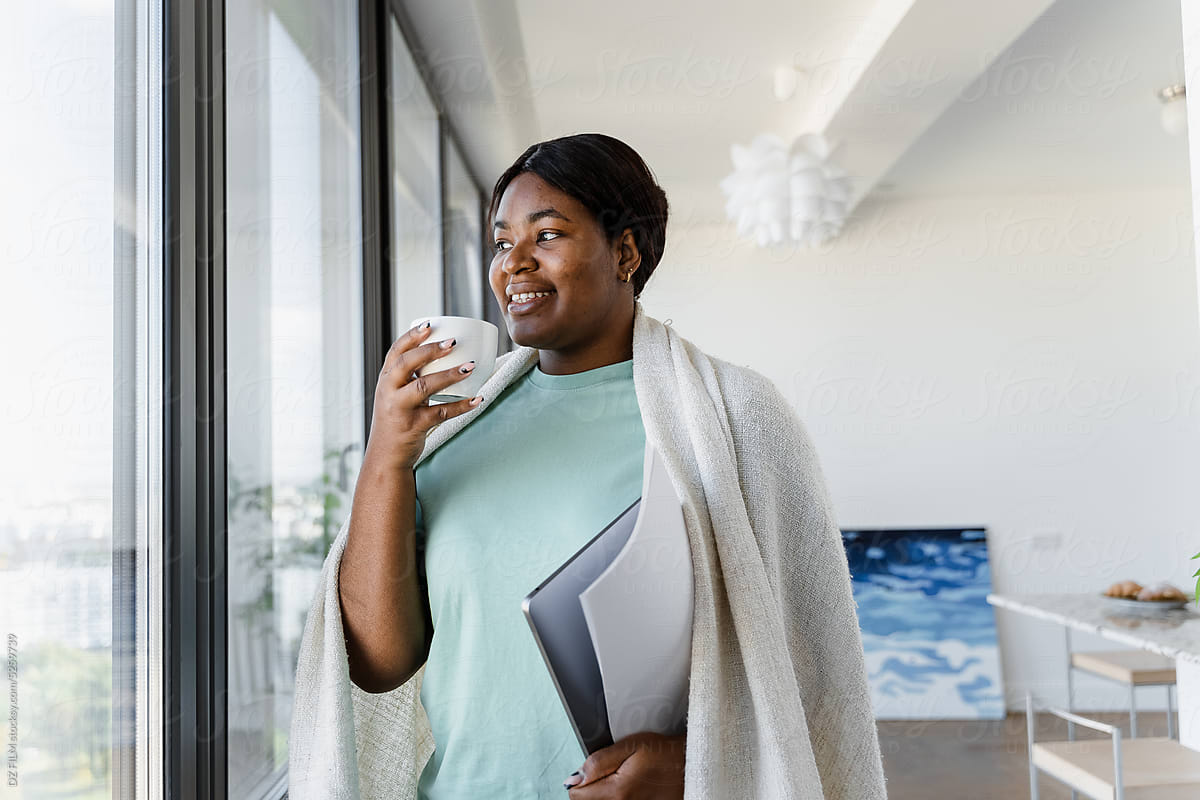 A woman with a laptop drinks coffee standing near the window