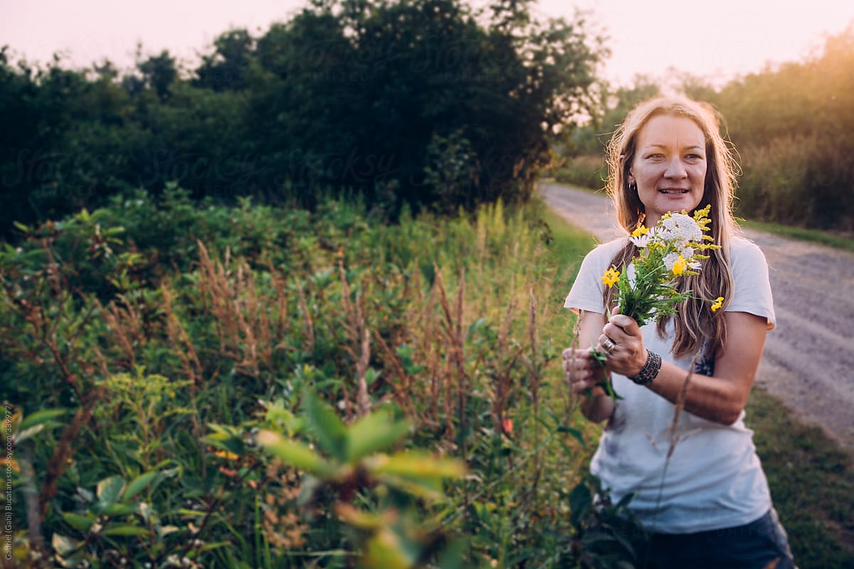 Woman Picking Wild Flowers By A Road Side At Sunset