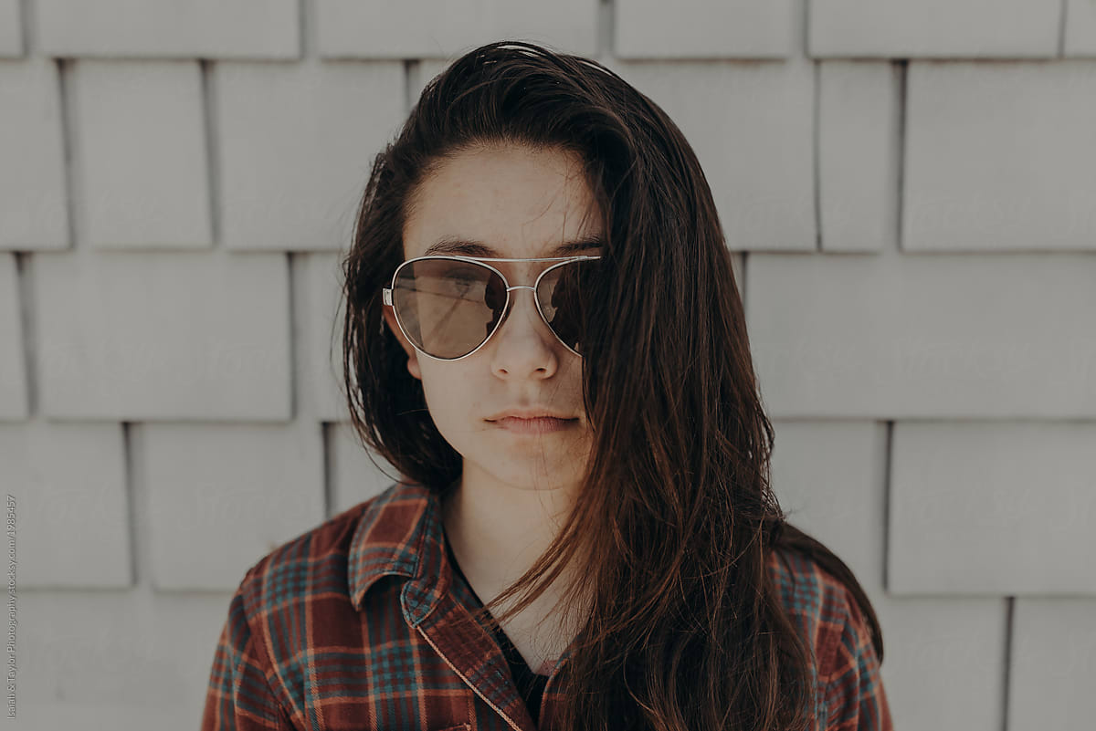 Portrait of young adult woman with long brown hair standing against a white textured wall staring straight at the camera with aviator sunglasses with a serious face