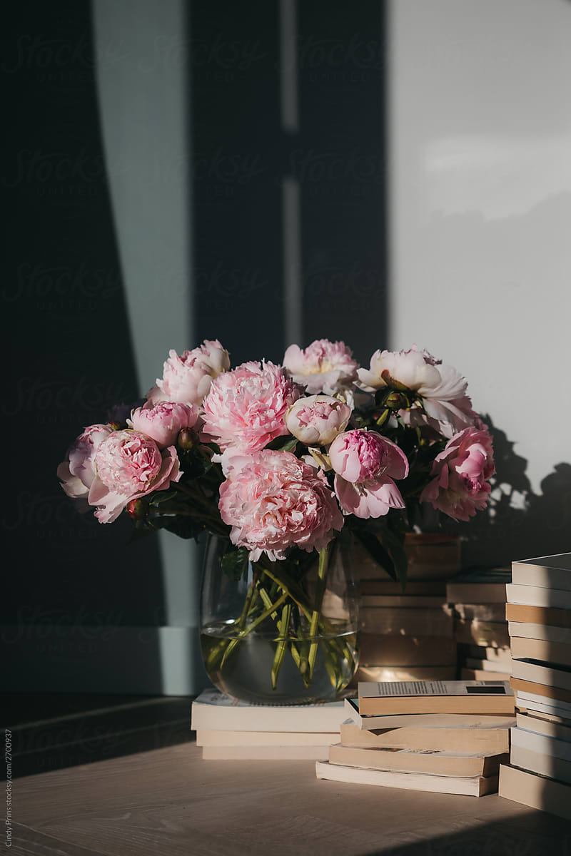 A glass vase of pink peonies on stacks of vintage books
