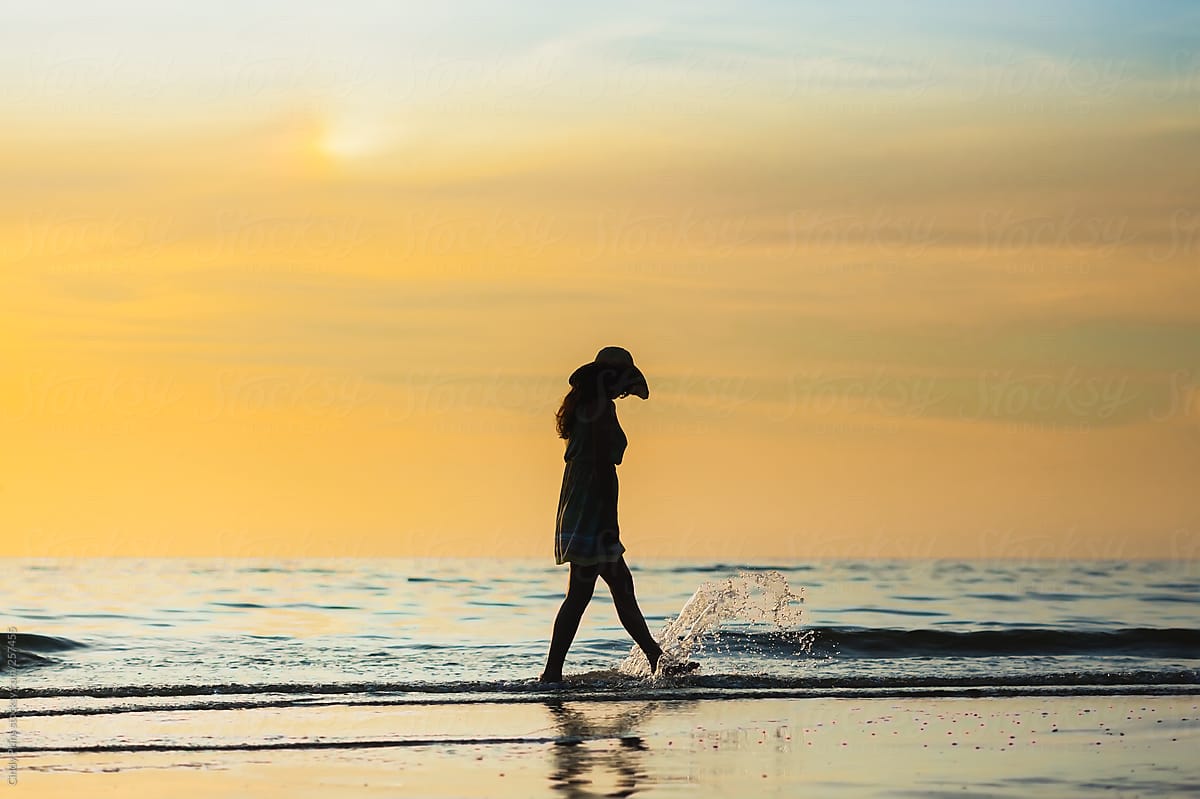 Silhouette Of A Woman With A Sunhat Walking Along The Shoreline At