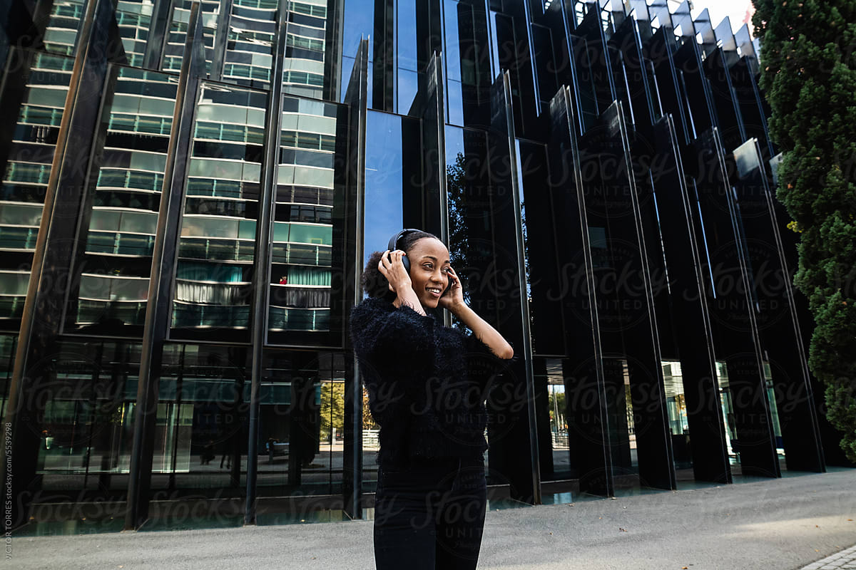 Cheerful black woman in headphones standing near city building