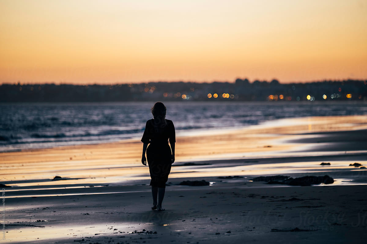 Barefooted lady walking along sandy seashore at sunset