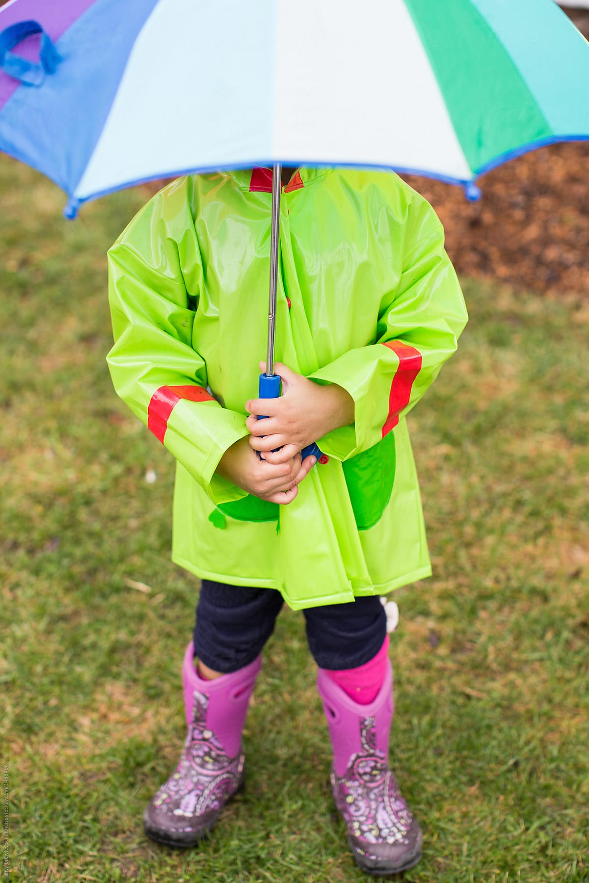 A young child in a raincoat holding a rainbow umbrella