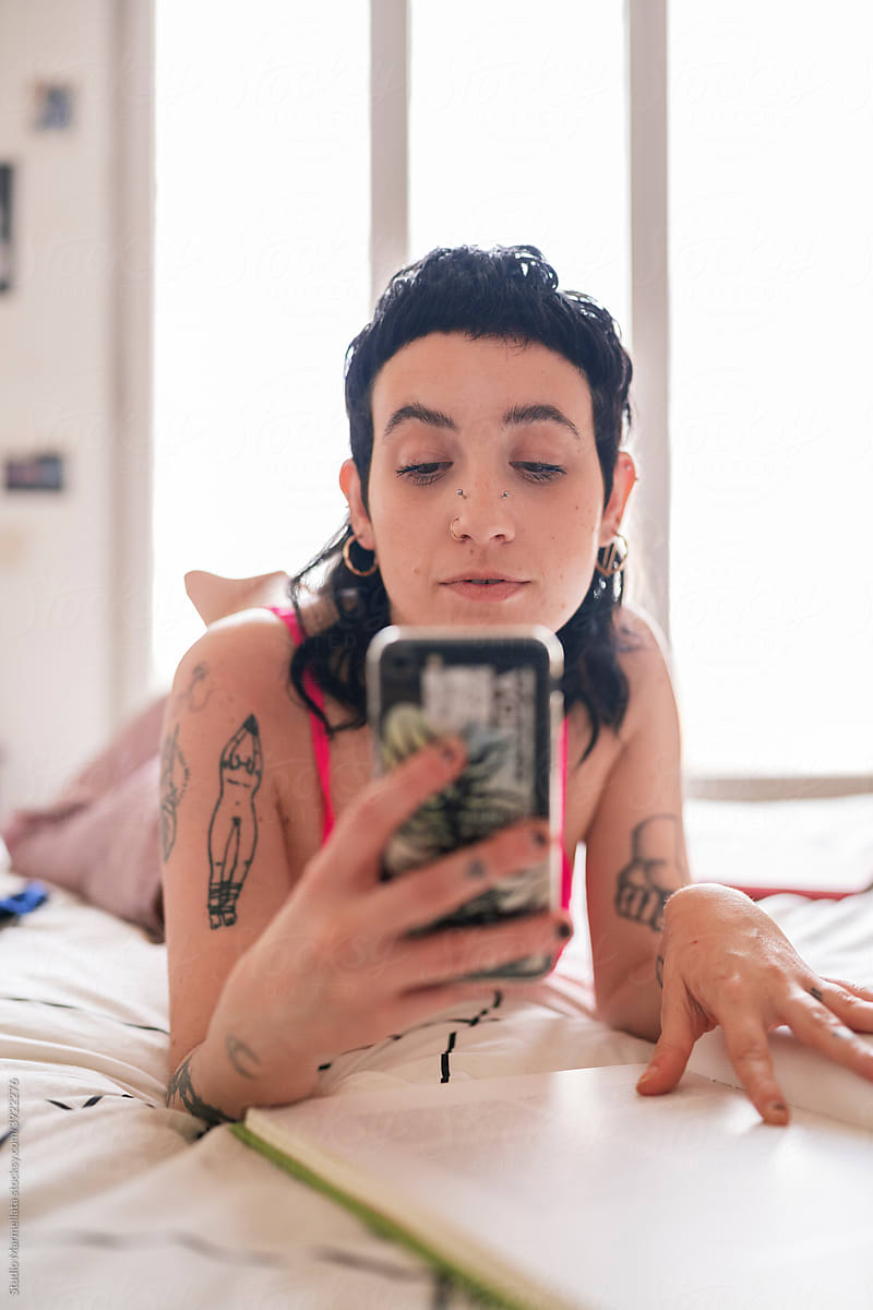 Tattooed woman using smartphone and reading book on bed