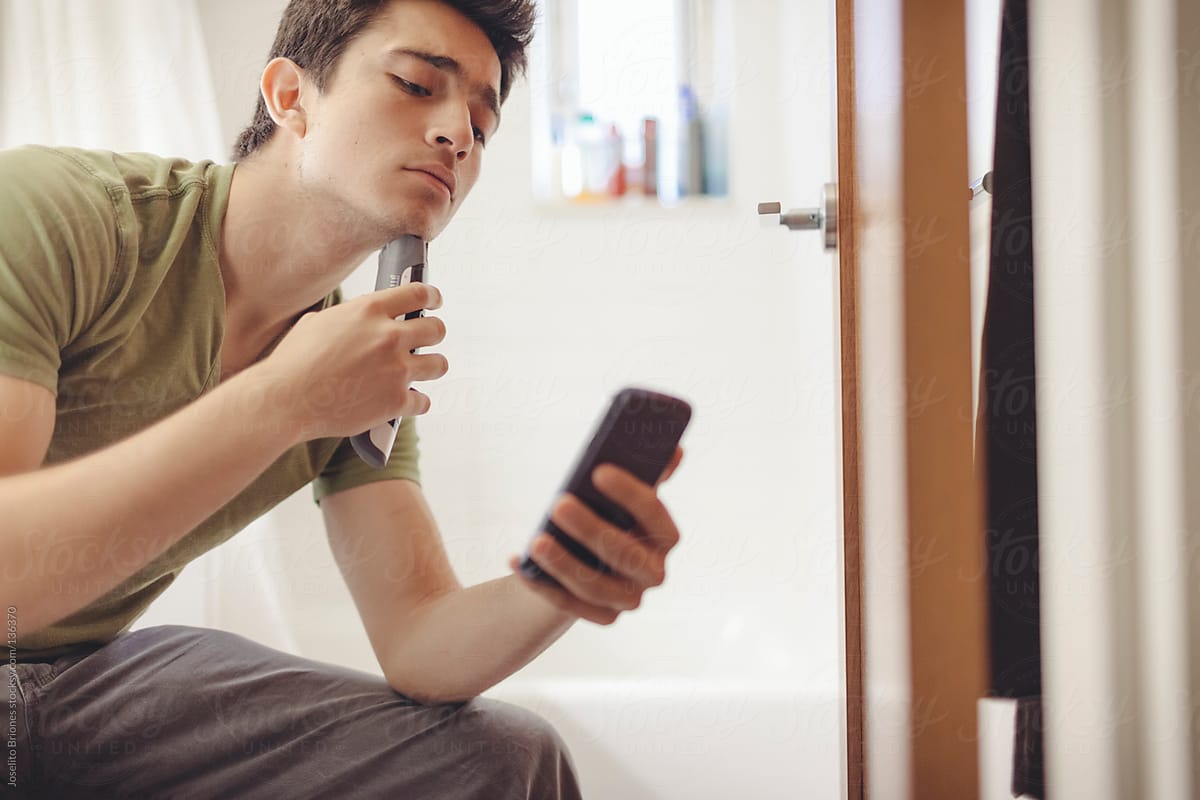 Young Mexican-American Man Shaving Using Smartphone Camera App as Mirror