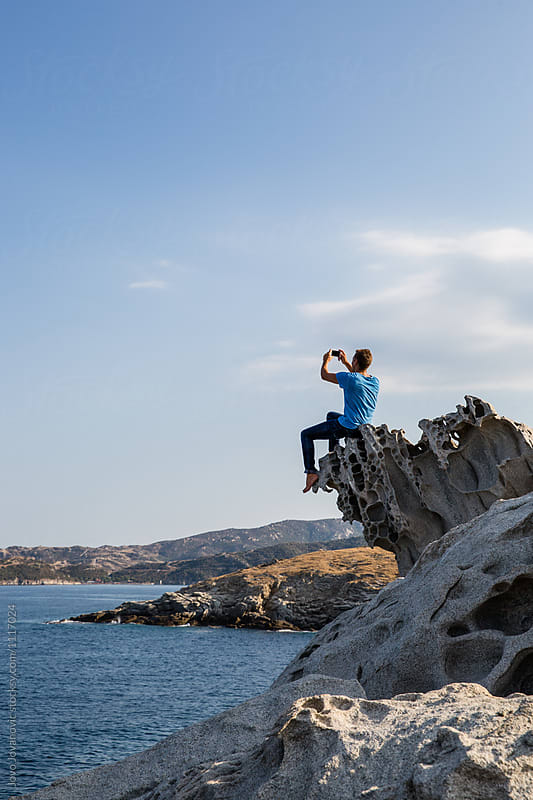 Man taking pictures of amazing landscape on smart phone camera while sitting on a rock near the sea