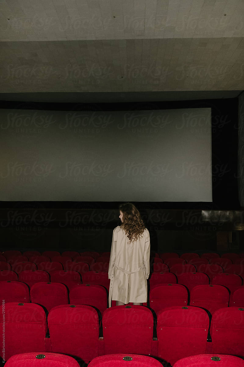 girl with curly hair stands between the rows of red cinema seats on the background of the screen