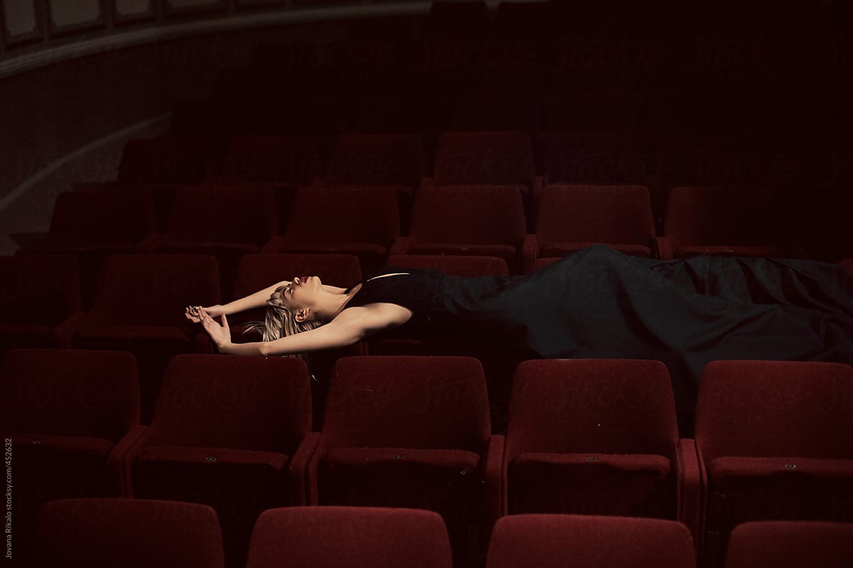 Young woman in a long black dress lies down on a theater chairs