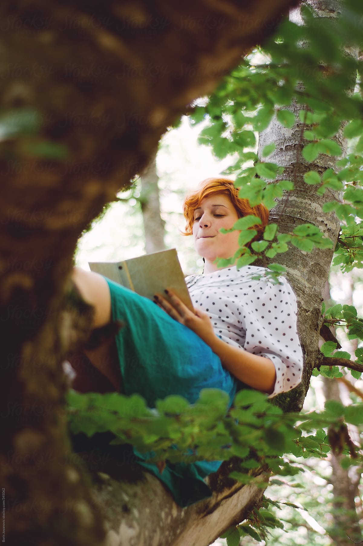 Young ginger girl laying on tree branch and reading a book