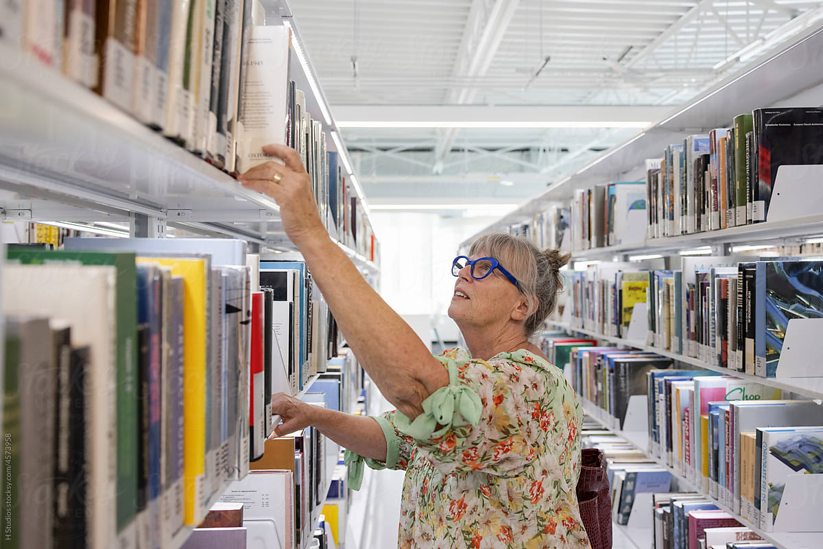 Senior woman reaching for book on library bookshelf.