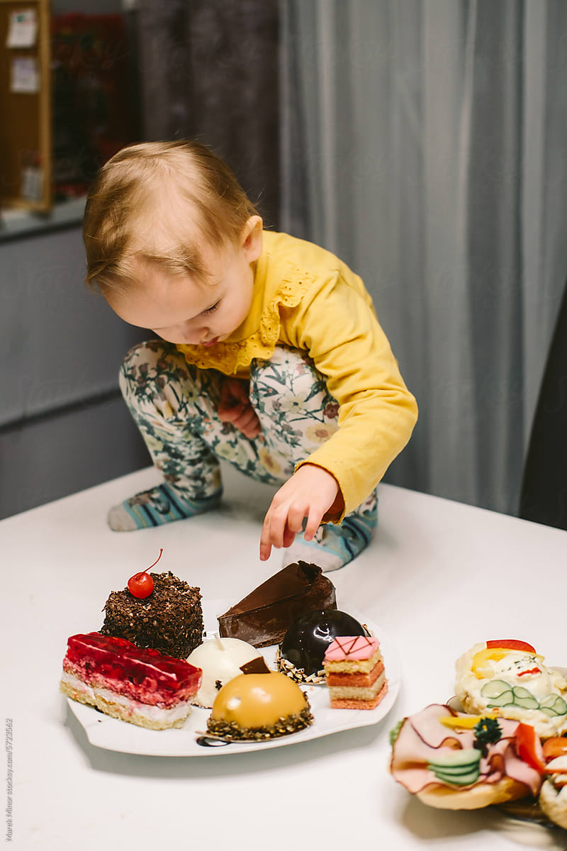 Small baby girl squats on a table, picking a cake from plate