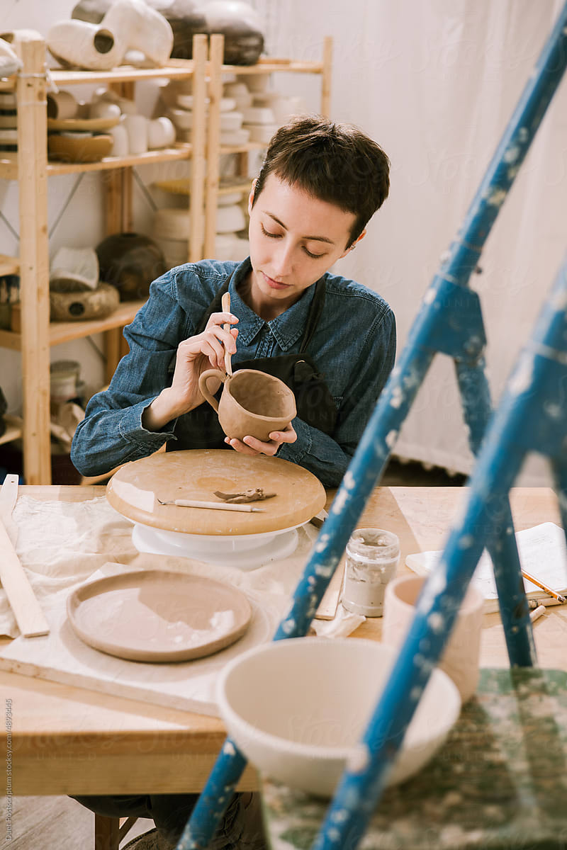 A young woman in an apron sculpts a mug at a work table in a workshop