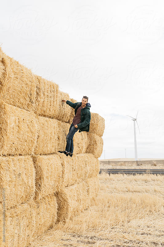Young adult male climbing on stacks of hay in recently plowed field