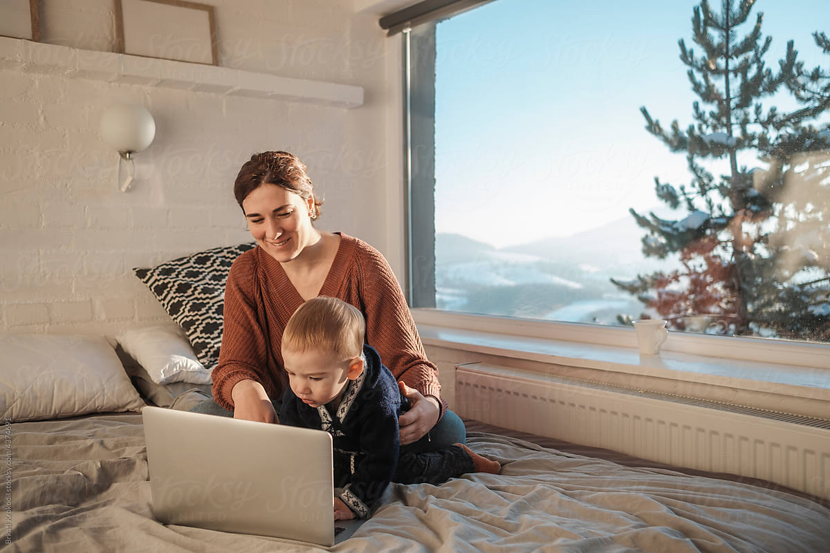 Smiling Mom And Baby Boy Sitting On The Bed With Laptop