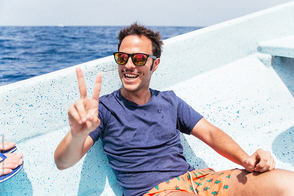 Fun portrait of a young man making the victory gesture with his hand on the edge of a boat with the water of the sea behind