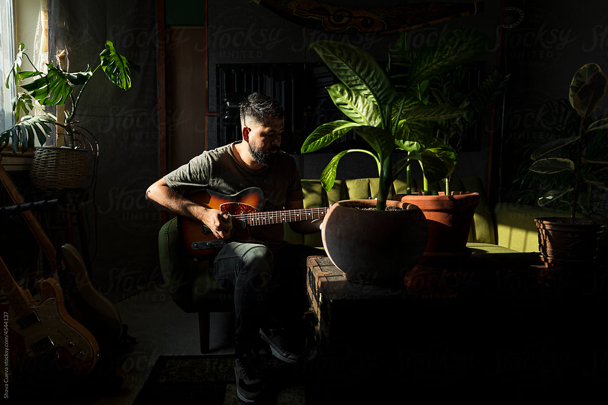 Man with beard playing an electric guitar in a  dark room with plants