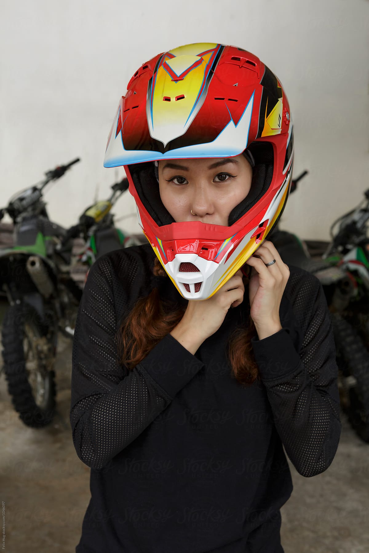 An asian woman is putting on the helmet for her motocross adventure. Behind her we some bikes.