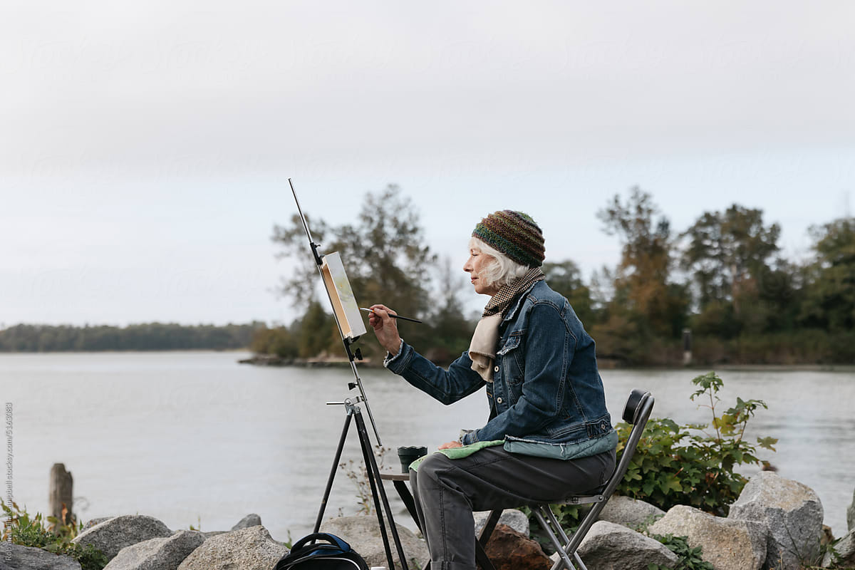 Silver haired woman painting on canvas by the river.