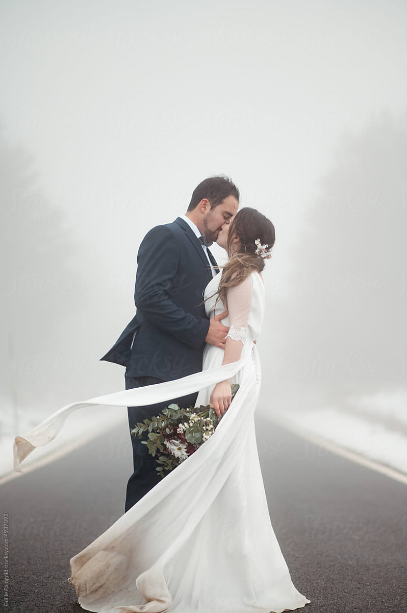 Newlywed kissing on road in wind.