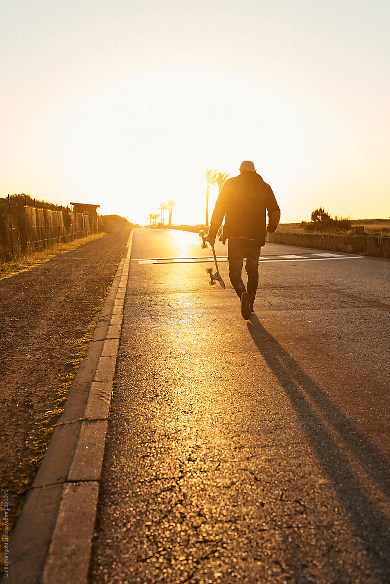 Man walking away with skateboard at sunset.