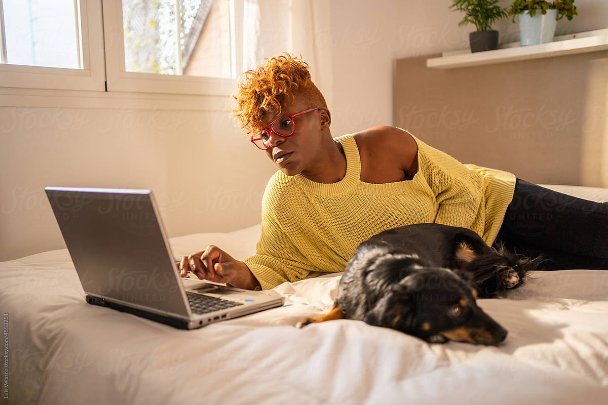 Black Woman With Laptop Next To Her Dog On The Bed.