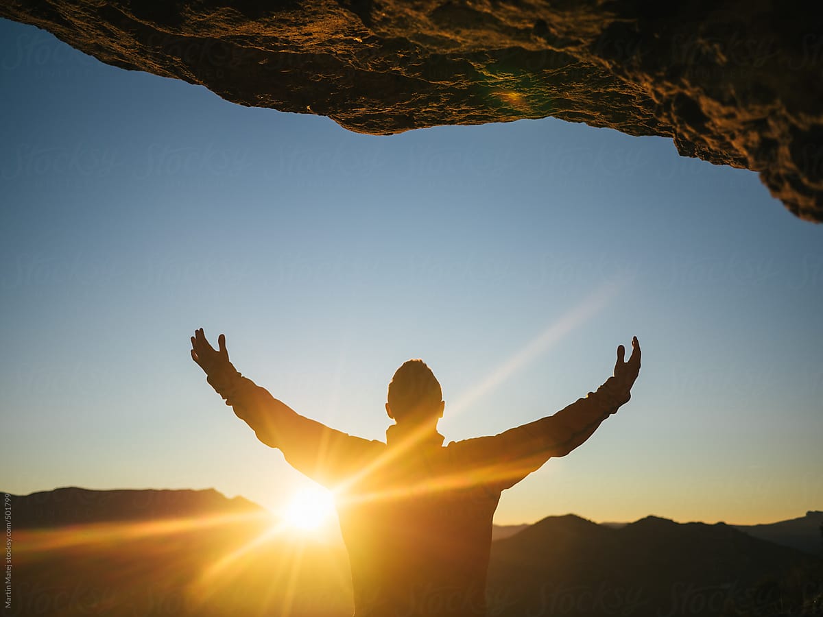 Man from behind with raised hands under a cliff by sunset