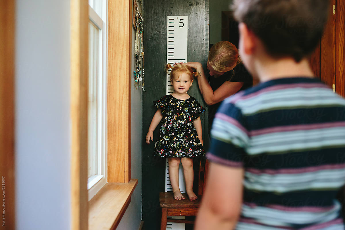 little girl stands on chair to be taller while mom measures her height