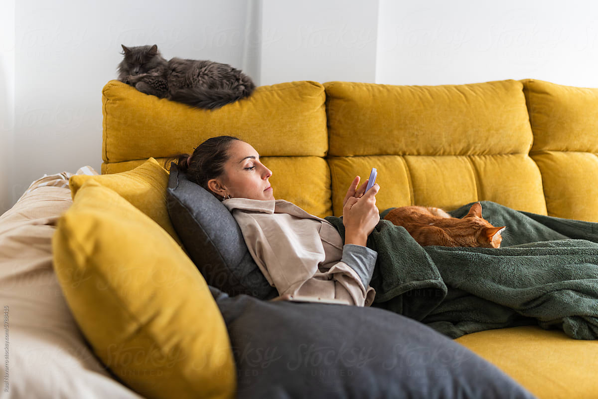 Beautiful woman lying on couch with cat and using smartphone at home